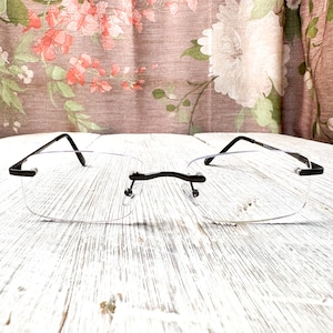 May include: Rimless eyeglasses with black temples and a black bridge. The lenses are clear and rectangular. The glasses are set against a white, textured wooden surface, with a floral patterned background.