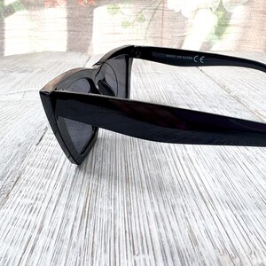 Puede incluir: Gafas de sol negras con forma de ojo de gato. Las gafas tienen un acabado brillante y lentes oscuras. Las patillas de las gafas son visibles y la montura es gruesa. Las gafas est&aacute;n sobre una superficie de madera clara.