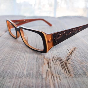 Può includere: Occhiali da vista rettangolari di colore marrone e arancio con un design decorativo sulle aste. Le lenti sono trasparenti.