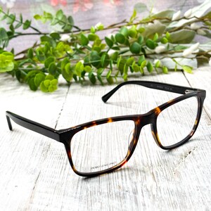 May include: Eyeglasses with a black and tortoiseshell pattern frame. The rectangular frames have clear lenses with the text "DEMO LENS ONLY". The glasses are set against a white wooden surface with green foliage in the background.