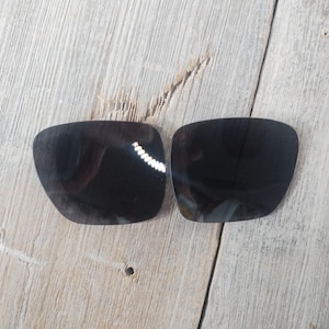 May include: Two black replacement lenses for sunglasses. The lenses are rectangular and have a slightly curved shape.