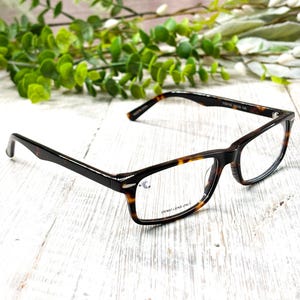 May include: Rectangular eyeglasses with a tortoise shell pattern on the frames. The lenses are clear, and the text "DEMO LENS ONLY" is visible on one lens. The glasses are set against a white wooden background with green foliage.