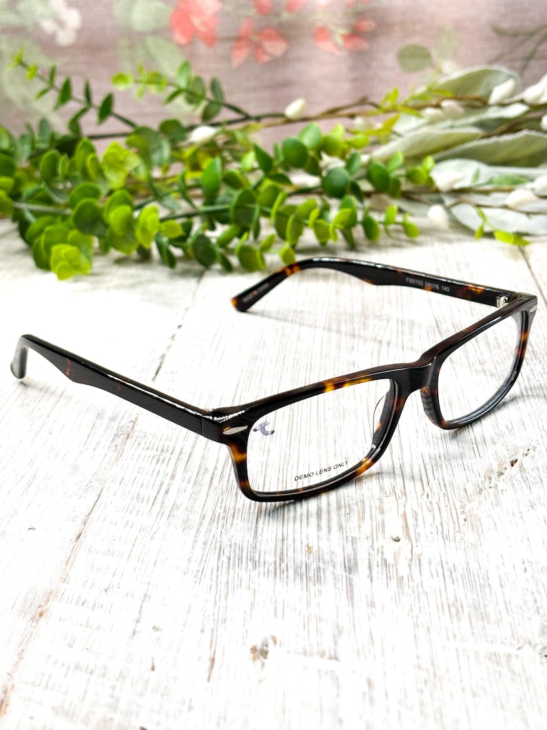 May include: Rectangular eyeglasses with a tortoise shell pattern on the frames. The lenses are clear, and the text "DEMO LENS ONLY" is visible on one lens. The glasses are set against a white wooden background with green foliage.