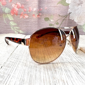May include: Brown gradient sunglasses with a gold metal frame and tortoise shell arms. The lenses transition from a dark brown at the top to a lighter brown at the bottom. The sunglasses are set against a floral background.