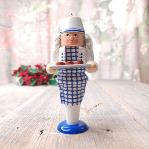 May include: A white and blue nutcracker figurine holding a tray with three brown pastries. The nutcracker is wearing a white hat and a blue and white checkered apron.