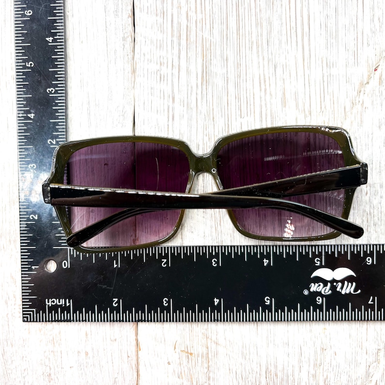 以下が含まれることがあります： 紫色のグラデーションレンズを備えた、濃いオリーブグリーンのサングラス。 サングラスは長方形のフレームと黒いアームを備えています。 画像には、サングラスの幅が約15cmであることを示す定規が含まれています。