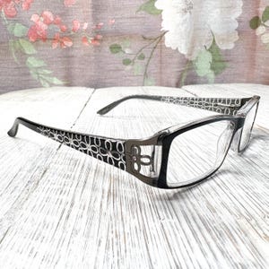May include: Black and silver eyeglasses with a floral design on the temples. The frames are rectangular with clear lenses. The glasses are set on a white wooden surface.