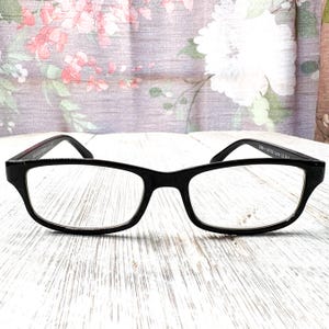 Könnte beinhalten: Schwarze rechteckige Brille mit klaren Gläsern. Die Fassungen sind dick und haben eine glänzende Oberfläche. Die Brille befindet sich vor einem floral gemusterten Hintergrund auf einer weißen Holzoberfläche.