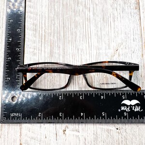 May include: Eyeglasses with a tortoise shell pattern frame, resting on a wooden surface. A black ruler with inch measurements is placed next to the glasses. The glasses have clear lenses and a rectangular shape.