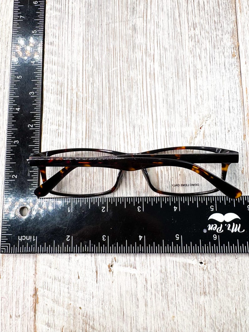 May include: Eyeglasses with a tortoise shell pattern frame, resting on a wooden surface. A black ruler with inch measurements is placed next to the glasses. The glasses have clear lenses and a rectangular shape.