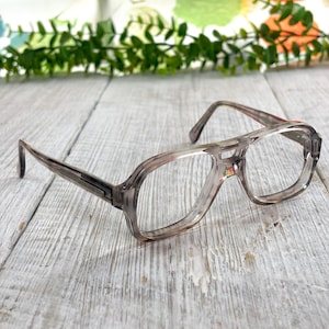 Puede incluir: Gafas de estilo aviador con una montura de acetato translúcido en color gris humo marmoleado. Las gafas presentan un diseño de doble puente, lentes rectangulares y varillas finas a juego. El material de la montura tiene un aspecto sutil, nublado y semitransparente.