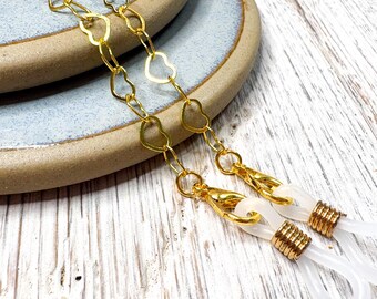 May include: Gold-colored eyeglass chain with heart-shaped links. The chain connects to white, rubberized loops that attach to the glasses. The chain is resting on a light blue and brown ceramic plate and a white wooden surface.