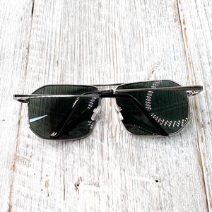 Pode incluir: &Oacute;culos de sol com arma&ccedil;&atilde;o de metal prateado e lentes escuras. Os &oacute;culos t&ecirc;m formato retangular com design de ponte dupla. As lentes s&atilde;o verde-escuras e a arma&ccedil;&atilde;o &eacute; prateada brilhante.