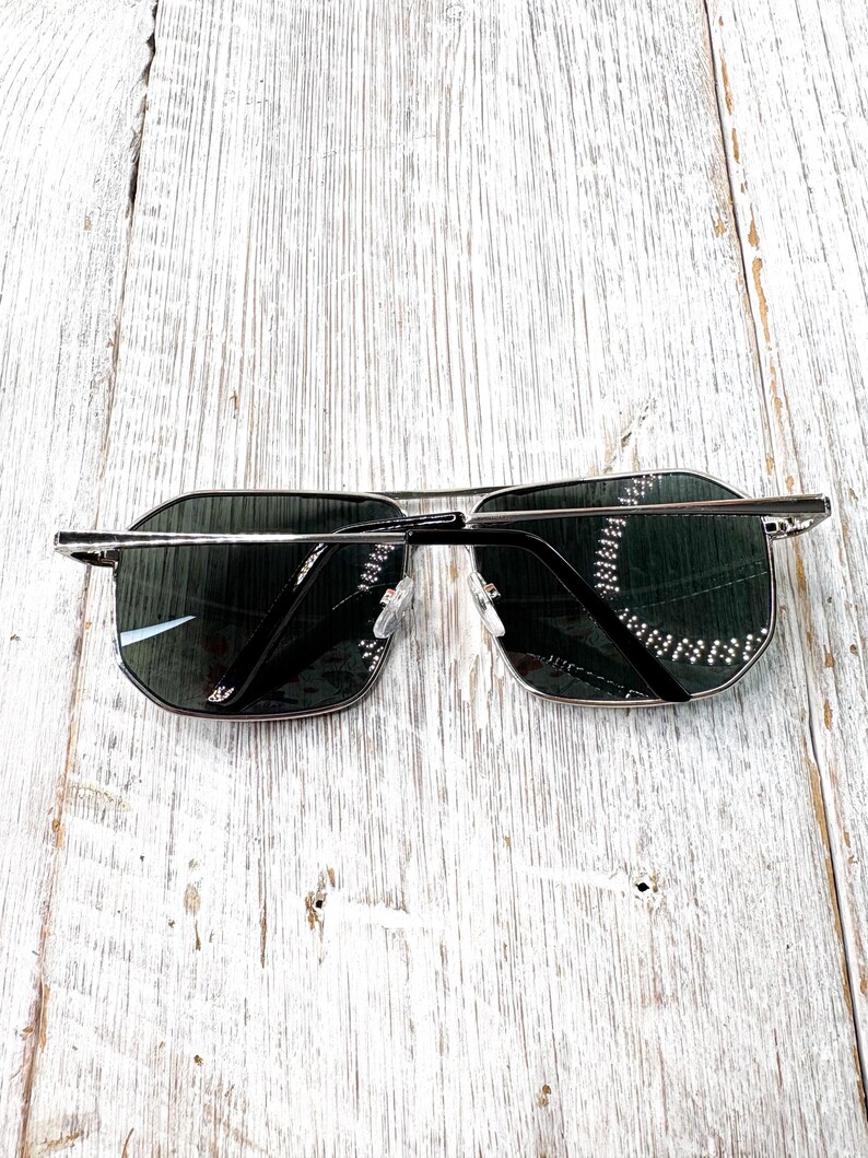 Pode incluir: &Oacute;culos de sol com arma&ccedil;&atilde;o de metal prateado e lentes escuras. Os &oacute;culos t&ecirc;m formato retangular com design de ponte dupla. As lentes s&atilde;o verde-escuras e a arma&ccedil;&atilde;o &eacute; prateada brilhante.