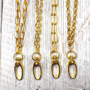 May include: Four gold-colored chain lanyards with lobster claw clasps. The chains have different link styles, including oval, rectangular, and round links. The clasps are attached to a ring at the top of each chain.