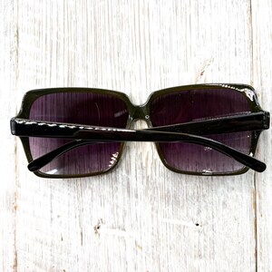 以下が含まれることがあります： 四角いフレームとグラデーションパープルのレンズを備えた、濃い緑色のサングラス。 サングラスは黒いブリッジとアームを備えています。 画像は真上から撮影されており、明るい色の木製の表面にサングラスが写っています。