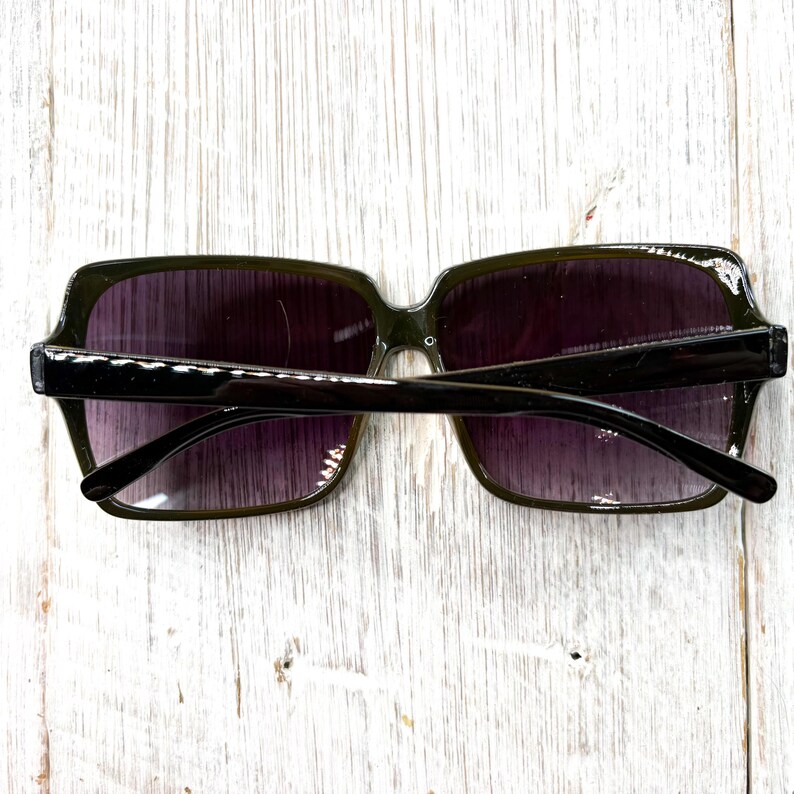 以下が含まれることがあります： 四角いフレームとグラデーションパープルのレンズを備えた、濃い緑色のサングラス。 サングラスは黒いブリッジとアームを備えています。 画像は真上から撮影されており、明るい色の木製の表面にサングラスが写っています。