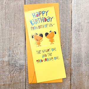 May include: A yellow birthday card with the text "HAPPY BIRTHDAY FROM BOTH OF US" and "THE SMART ONE AND THE GOOD-LOOKING ONE." Two cartoon dogs are featured on the card, which is inside a matching envelope.