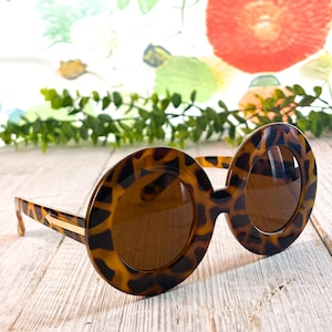 May include: Round sunglasses featuring a brown and black tortoiseshell pattern on the frames and temples. The lenses are tinted a solid brown color. A thin, horizontal gold-toned metal accent is visible on the side of the temple near the hinge.