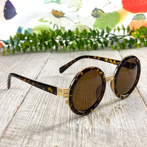 May include: Round sunglasses featuring a tortoiseshell pattern on the frames and temples, accented with gold-tone metal detailing at the hinges and bridge. The lenses are a solid brown tint, providing a classic look with a circular frame shape.