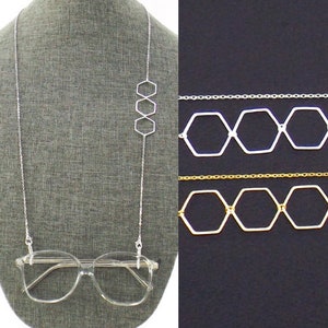 Op de afbeelding: Een zilveren ketting met drie zeshoekige bedels hangt aan een transparant plastic montuur voor een bril. De ketting wordt getoond tegen een grijze achtergrond. De ketting wordt ook getoond tegen een zwarte achtergrond met twee andere kettingen, een zilveren en een gouden, met zeshoekige bedels.