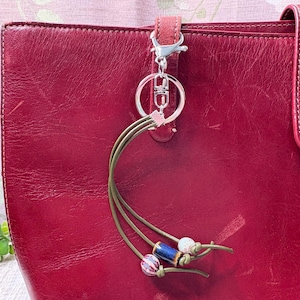 May include: A red leather tote bag with a silver keyring and a decorative tassel. The tassel features olive green cords and decorative beads in white, blue, and pink. The bag has a matching red strap.