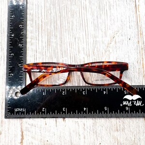 Puede incluir: Gafas con estampado de carey, con montura rectangular. Las gafas se miden con una regla, mostrando una longitud de aproximadamente 13 cm. Las lentes son transparentes y la montura tiene un dise&ntilde;o cl&aacute;sico.