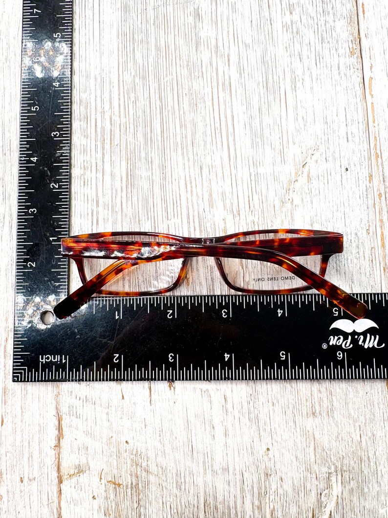 Puede incluir: Gafas con estampado de carey, con montura rectangular. Las gafas se miden con una regla, mostrando una longitud de aproximadamente 13 cm. Las lentes son transparentes y la montura tiene un dise&ntilde;o cl&aacute;sico.