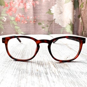 Puede incluir: Gafas de montura redonda con estampado de carey. Las gafas tienen lentes transparentes y están colocadas sobre una superficie de madera blanca con un fondo floral.