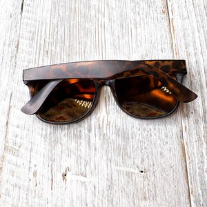 May include: Tortoiseshell sunglasses with dark lenses. The sunglasses have a classic rectangular frame and are set against a light-colored, weathered wood background. The arms of the sunglasses are slightly open.