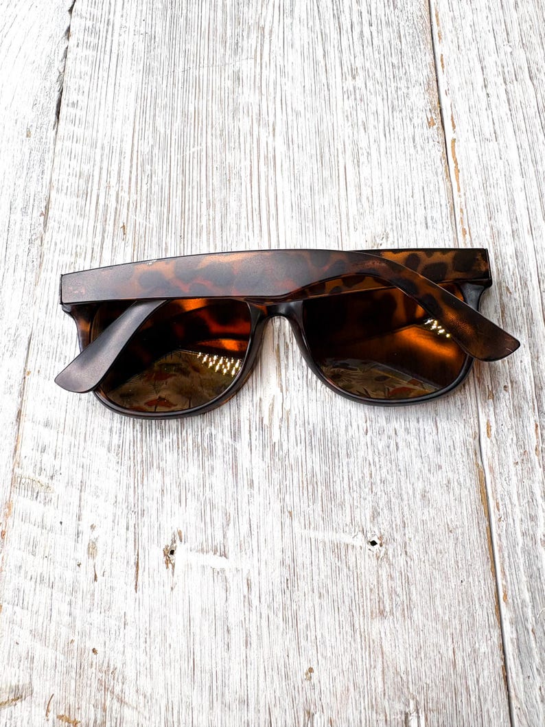 May include: Tortoiseshell sunglasses with dark lenses. The sunglasses have a classic rectangular frame and are set against a light-colored, weathered wood background. The arms of the sunglasses are slightly open.