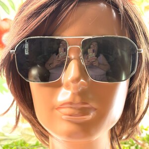 Pode incluir: &Oacute;culos de sol com arma&ccedil;&atilde;o prateada e lentes escuras expostos em uma cabe&ccedil;a de manequim com cabelo castanho. Os &oacute;culos t&ecirc;m formato retangular com ponte dupla. As lentes refletem o ambiente.