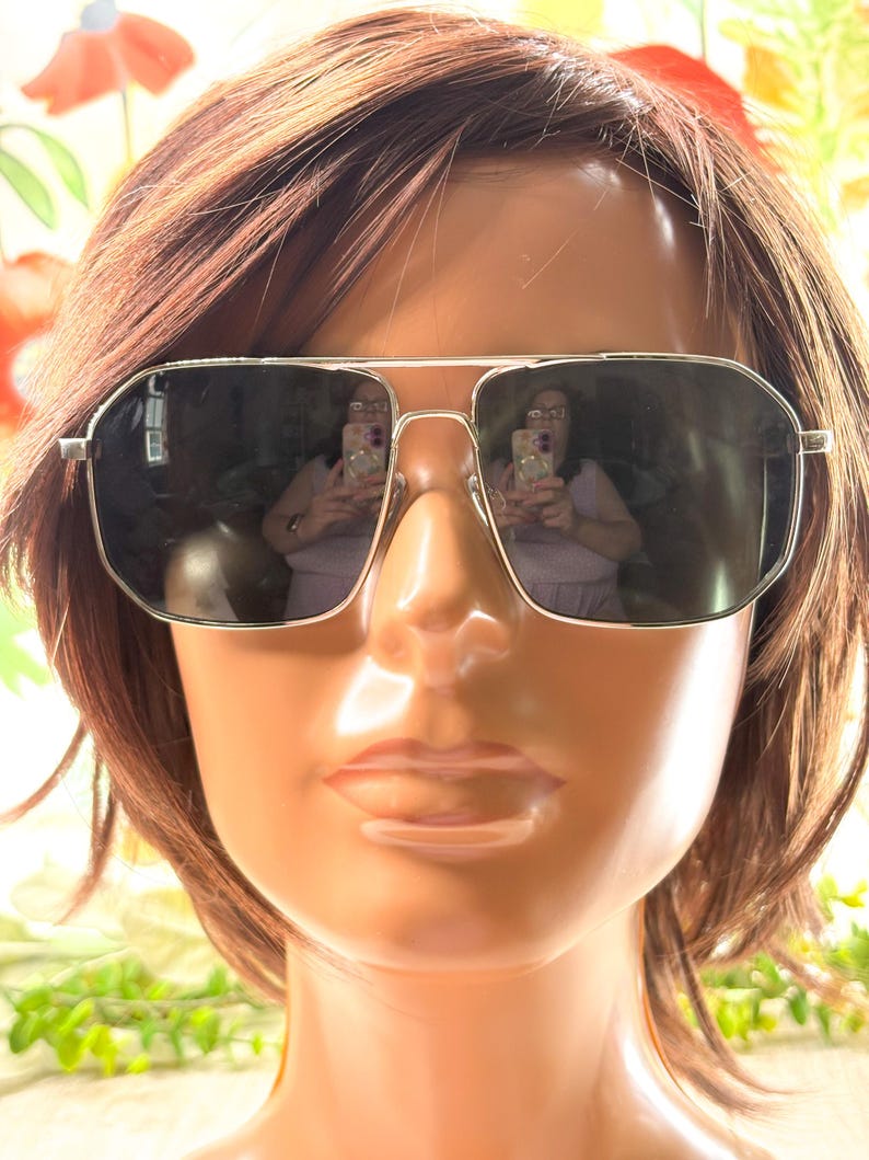 Pode incluir: &Oacute;culos de sol com arma&ccedil;&atilde;o prateada e lentes escuras expostos em uma cabe&ccedil;a de manequim com cabelo castanho. Os &oacute;culos t&ecirc;m formato retangular com ponte dupla. As lentes refletem o ambiente.