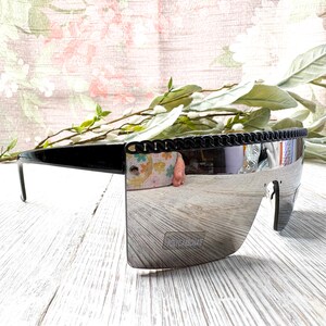 Può includere: Occhiali da sole neri con lente a specchio. Gli occhiali hanno una spessa montatura nera con un dettaglio a catena lungo la parte superiore. La lente è di colore argento riflettente. Il testo "POLYCARBONATE" è stampato sulla lente.