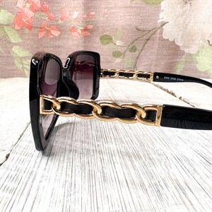Puede incluir: Gafas de sol negras con dise&ntilde;o de cadena dorada en las patillas. Las lentes son oscuras y las monturas cuadradas. Las gafas de sol est&aacute;n sobre una superficie de madera blanca. El texto "SWG 2008 China" est&aacute; impreso en la patilla.