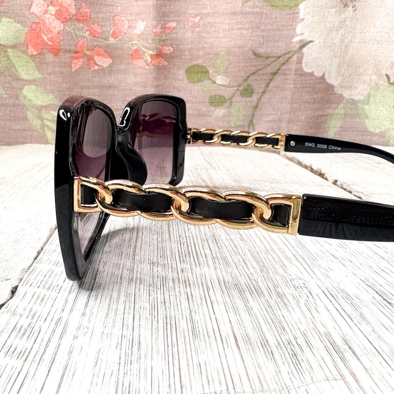 Puede incluir: Gafas de sol negras con dise&ntilde;o de cadena dorada en las patillas. Las lentes son oscuras y las monturas cuadradas. Las gafas de sol est&aacute;n sobre una superficie de madera blanca. El texto "SWG 2008 China" est&aacute; impreso en la patilla.