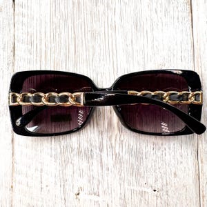 Puede incluir: Gafas de sol negras con detalles de cadena dorada en las patillas. Las lentes tienen un degradado, oscuro en la parte superior y m&aacute;s claro en la inferior. La montura es rectangular y brillante.