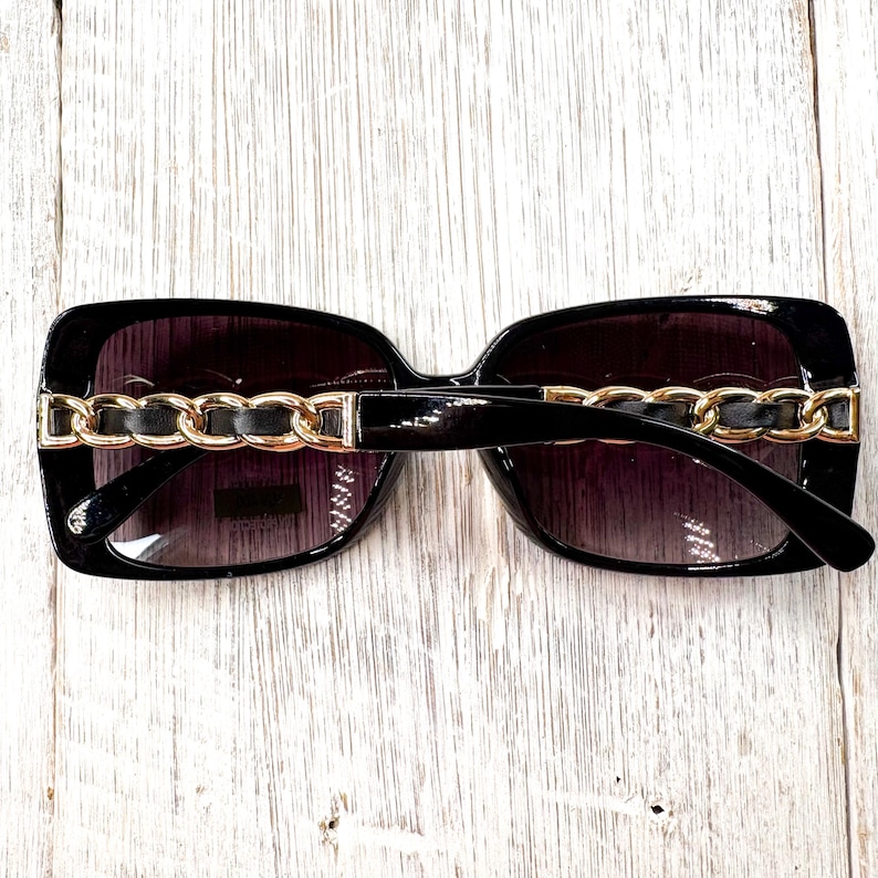 Puede incluir: Gafas de sol negras con detalles de cadena dorada en las patillas. Las lentes tienen un degradado, oscuro en la parte superior y m&aacute;s claro en la inferior. La montura es rectangular y brillante.