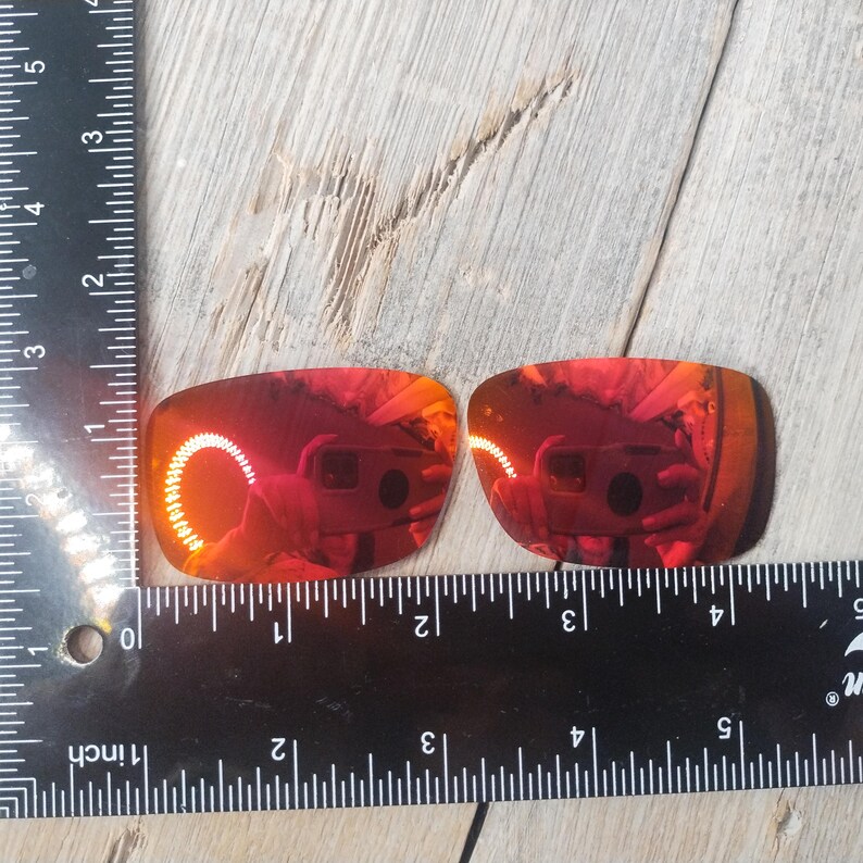 Może przedstawiać: Dwa czerwone, lustrzane szkła zamienne do okular&oacute;w przeciwsłonecznych, zmierzone linijką. Szkła mają około 5 cm szerokości.