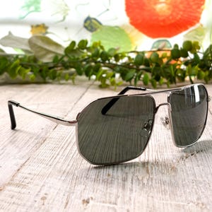 Pode incluir: &Oacute;culos de sol aviador prateados com lentes escuras. Os &oacute;culos t&ecirc;m um design cl&aacute;ssico de dupla ponte e um visual elegante e moderno. As lentes s&atilde;o de um tom verde escuro e as arma&ccedil;&otilde;es s&atilde;o prateadas brilhantes.