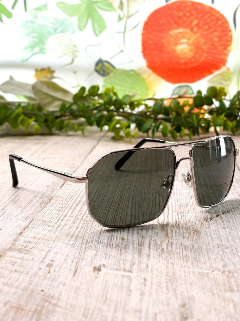 Pode incluir: &Oacute;culos de sol aviador prateados com lentes escuras. Os &oacute;culos t&ecirc;m um design cl&aacute;ssico de dupla ponte e um visual elegante e moderno. As lentes s&atilde;o de um tom verde escuro e as arma&ccedil;&otilde;es s&atilde;o prateadas brilhantes.