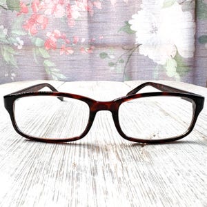 May include: A pair of eyeglasses with a tortoiseshell pattern on the frames. The rectangular lenses are clear, and the glasses are set against a floral background. The frames have a dark brown and black color.