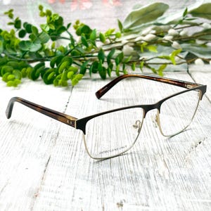 May include: Eyeglasses with a black and gold frame. The temples are a tortoiseshell pattern. The text "DEMO LENS ONLY" is printed on the lens. The glasses are set against a white wooden background with green foliage.