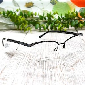 May include: Black and silver half-rim eyeglasses with a patterned design on the upper frame. The lenses are clear, and the text "DEMO LENS ONLY" is visible. The glasses rest on a white wooden surface.