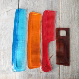 May include: Four combs of different colors and styles are arranged on a light wood surface. The combs include blue, orange, red, and brown. The red comb has a handle. The combs are made of plastic and wood.