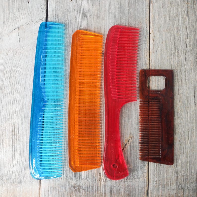 May include: Four combs of different colors and styles are arranged on a light wood surface. The combs include blue, orange, red, and brown. The red comb has a handle. The combs are made of plastic and wood.