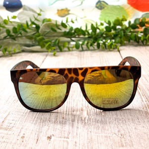 May include: Brown tortoiseshell sunglasses with mirrored yellow lenses. The sunglasses have a rounded frame shape and are set against a light-colored wooden surface. The lenses have UV 400 protection.