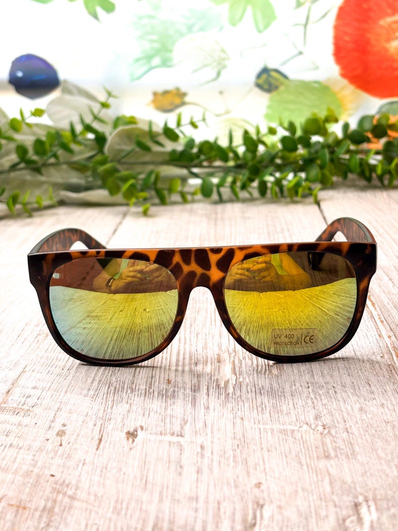 May include: Brown tortoiseshell sunglasses with mirrored yellow lenses. The sunglasses have a rounded frame shape and are set against a light-colored wooden surface. The lenses have UV 400 protection.
