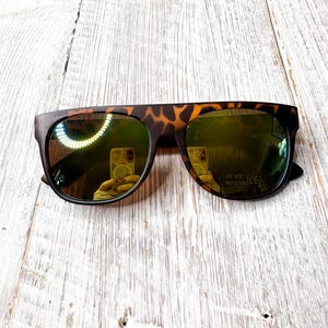 May include: Sunglasses with a tortoiseshell pattern on the frame and dark, reflective lenses. The sunglasses have a flat top design and are set against a light-colored wooden surface. The lenses have a UV 400 protection label.
