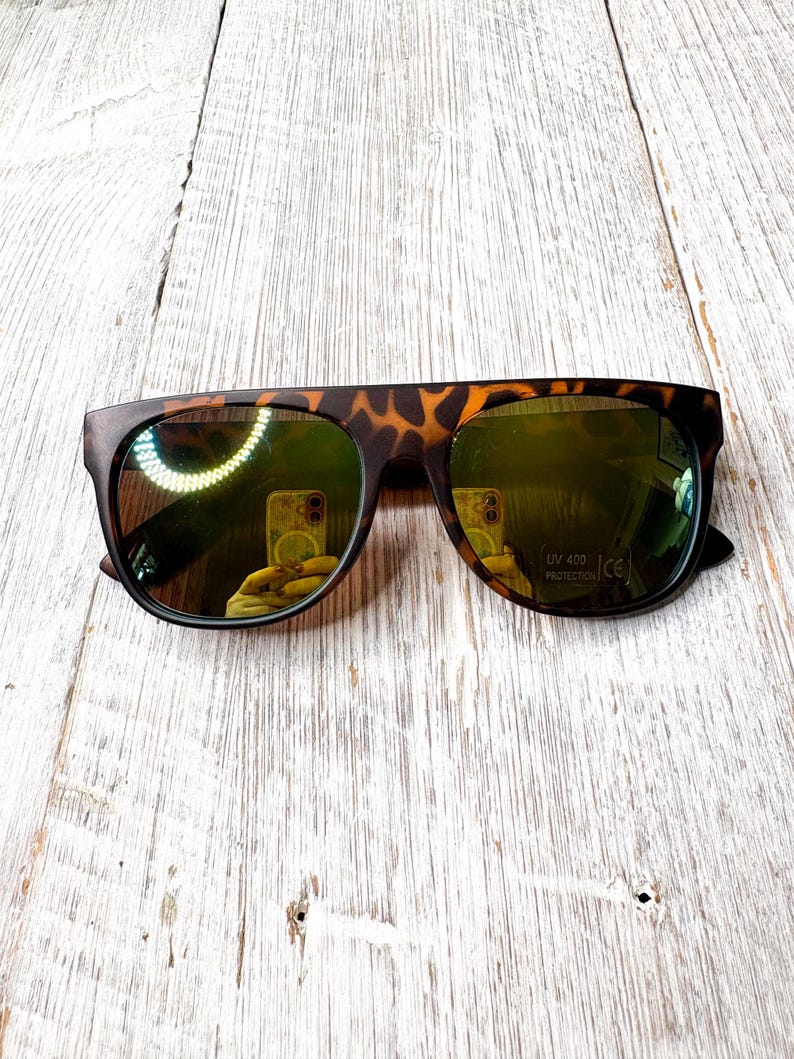 May include: Sunglasses with a tortoiseshell pattern on the frame and dark, reflective lenses. The sunglasses have a flat top design and are set against a light-colored wooden surface. The lenses have a UV 400 protection label.